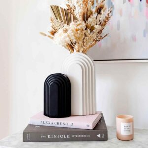 1 white ceramic vase with gold artificial flowers and 1 small black vase at the table with two books and 1 decoration piece.