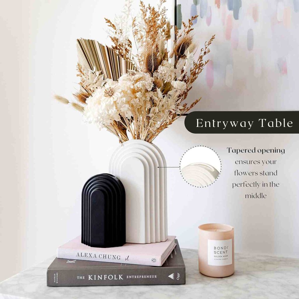 1 white ceramic vase with gold artificial flowers and 1 small black vase at the table with two books and 1 decoration piece. 